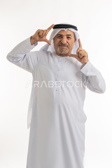 مفهوم الرجولة والثقة بالنفس، بورتريه لرجل مسن عربي خليجي إماراتي يرتي الكندورة والغترة ينظر الى الكاميرا بايماءات البشاشة والسرور، تعابير وحركات يدين تدل على التقاط صور، توثيق اللحظات السعيدة, الاهتمام بالأناقة والمظهر الخارجي، خلفية بيضاء