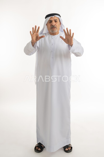 الهلع وطلب التوقف وترك مسافة أمان، الذعر والخوف من شيء ما، مد اليدين نحو الأمام في محاولة للدفاع عن النفس، بورتريه لرجل مسن عربي خليجي إماراتي يرتدي الكندورة والغترة ينظر إلى الكاميرا بإيماءات الرعب والرهبة، الجسم بالطول الكامل، خلفية بيضاء