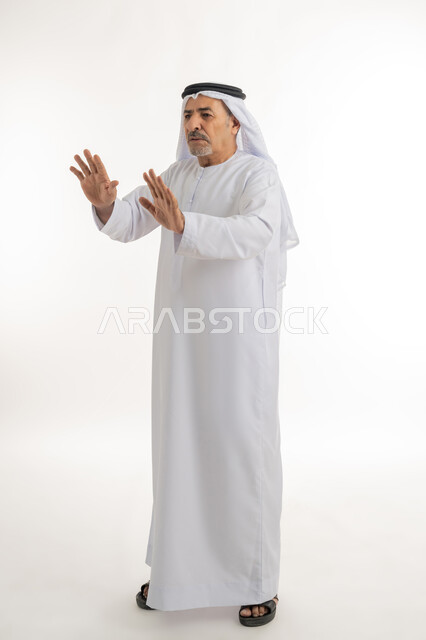 ترك مسافة أمان وطلب التوقف، الذعر والخوف من شيء ما، مد اليدين نحو الأمام في محاولة للدفاع عن النفس، بورتريه لرجل مسن عربي خليجي إماراتي يرتدي الكندورة والغترة ينظر إلى الكاميرا بإيماءات الرعب والرهبة، الجسم بالطول الكامل، خلفية بيضاء