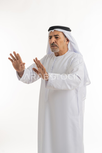 الذعر والخوف من شيء ما، مد اليدين نحو الأمام في محاولة للدفاع عن النفس، بورتريه لرجل مسن عربي خليجي إماراتي يرتدي الكندورة والغترة ينظر إلى الكاميرا بإيماءات الرعب والرهبة، ترك مسافة أمان وطلب التوقف، خلفية بيضاء