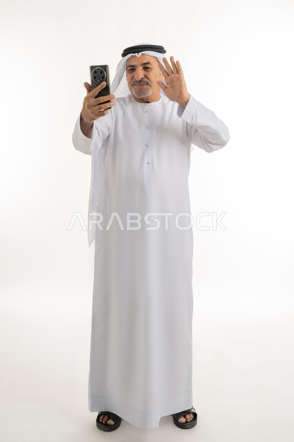 القاء التحية من خلال شاشة الموبايل، اجراء مكالمة فيديو عبر جهاز الجوال، بورتريه لرجل عربي خليجي اماراتي يرتدي الكندورة والغترة يقوم باستخدام الهاتف المحمول، التواصل مع الأهل والأصدقاء وسماع أخبارهم، الطول الكامل للجسم، خلفية بيضاء
