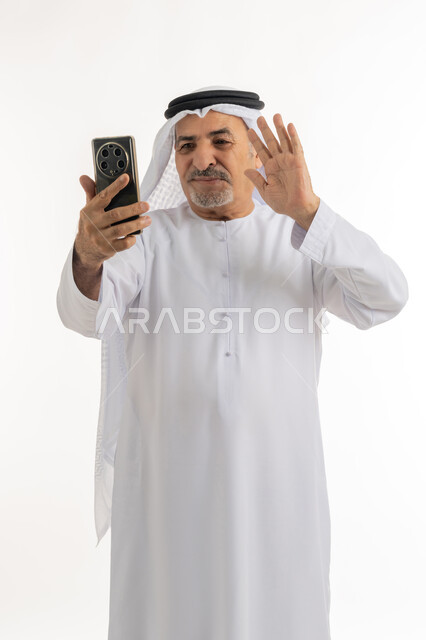اجراء مكالمة فيديو عبر جهاز الجوال، بورتريه لرجل عربي خليجي اماراتي يرتدي الكندورة والغترة يقوم باستخدام الهاتف المحمول، التواصل مع الأهل والأصدقاء وسماع أخبارهم، القاء التحية من خلال شاشة الموبايل، خلفية بيضاء