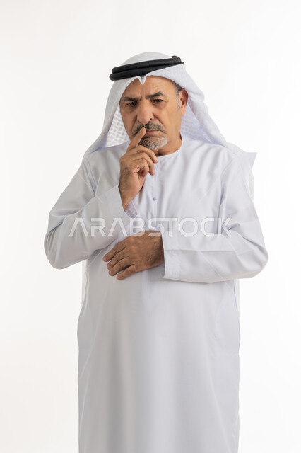 شرود الذهن و التمعن، النظر الى شيء ما بتعابير القلق،  بورتريه لرجل كبير بالسن عربي خليجي اماراتي يرتدي الكندورة والغترة يقف ويضع يده على ذقنه بإيماءات والتفكير بشيء ما, التخطيط ووضع الأهداف المستقبلية, خلفية بيضاء