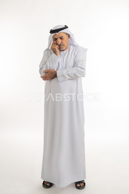 إيماءات التفكير العميق والحيرة، وضع اليد تحت الذقن بتعابير القلق وعدم اتخاذ القرار، بورتريه لرجل عربي خليجي إماراتي كبير بالسن يرتدي الكندورة الإماراتية التقليدية والغترة يقف وينظر إلى شيء ما ويخطط لشيء ما، الجسم بالطول الكامل، خلفية بيضاء