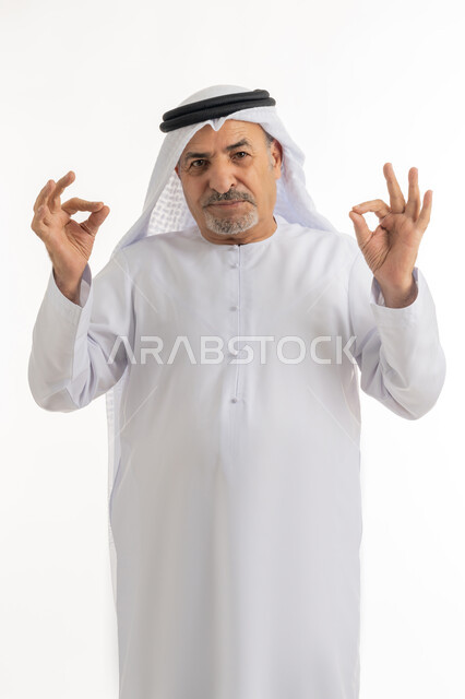 النظر للكاميرا بتعابير السعادة والرضا، بورتريه  لرجل عربي خليجي إماراتي كبير بالسن يرتدي الكندورة الإماراتية والغترة يقف ويرفع يديه بإيماءات الموافقة، إشارات يدين تدل على علامة الجودة والإعجاب، خلفية بيضاء