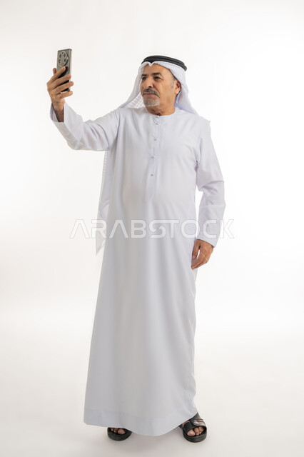 توثيق اللحظات السعيدة، بورتريه لرجل مسن عربي خليجي إماراتي يرتدي الكندورة والغترة يقوم بالتقاط صور السيلفي التذكارية عبر الهاتف المحمول بإيماءات السرور، استخدام التقنيات الحديثة المتطورة، صورة لكامل الجسم، خلفية بيضاء