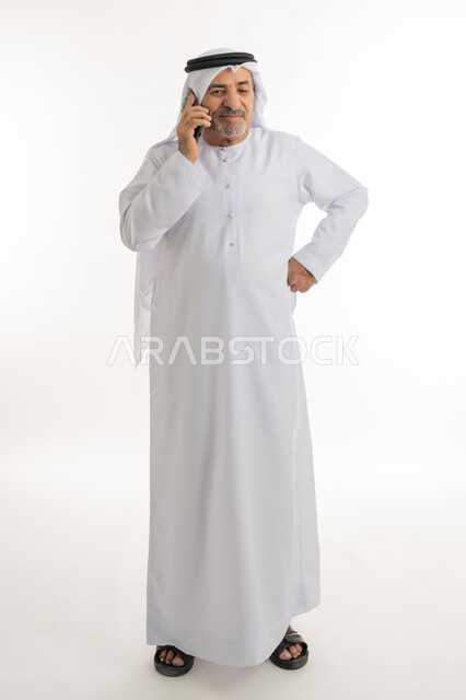 إيماءات وتعابير السعادة والسرور لسماع الأخبار المفرحة، بورتريه لرجل مسن عربي خليجي إماراتي مبتسم يرتدي الكندورة والغترة يقوم بإجراء مكالمة هاتفية عبر الهاتف المحمول، استخدام التقنيات والأجهزة الحديثة، التواصل مع الأهل والأصدقاء، الطول الكامل للجسم، خلفية بيضاء