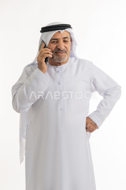 تبادل الأحاديث و الأخبار الجيدة،  بورتريه لرجل مسن عربي خليجي إماراتي مبتسم يرتدي الكندورة والغترة يقوم بإجراء مكالمة هاتفية عبر الهاتف المحمول،  التواصل مع الأهل والأصدقاء، ايماءات السعادة والسرور، خلفية بيضاء