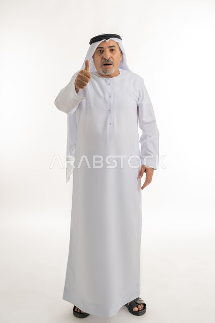 الاهتمام بالأناقة والمظهر الخارجي، بورتريه لرجل كبير بالسن عربي خليجي إماراتي يرتدي الكندورة والغترة ينظر الى الكاميرا يرفع إصبع السبابة للاعلى بإيماءات الإعجاب والرضا، تعابير القبول والذهول والصدمة، صورة الجسم بالطول الكامل، خلفية بيضاء
