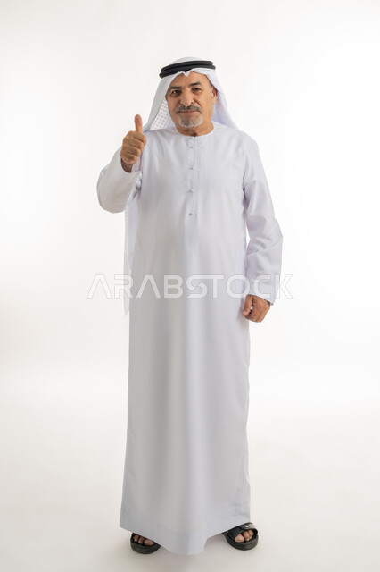تعابير القبول والفرح والسرور، الاهتمام بالأناقة والمظهر الخارجي، بورتريه لرجل كبير بالسن عربي خليجي إماراتي يرتدي الكندورة والغترة ينظر الى الكاميرا يرفع إصبع السبابة للاعلى بإيماءات الإعجاب والرضا، صورة الجسم بالطول الكامل، خلفية بيضاء
