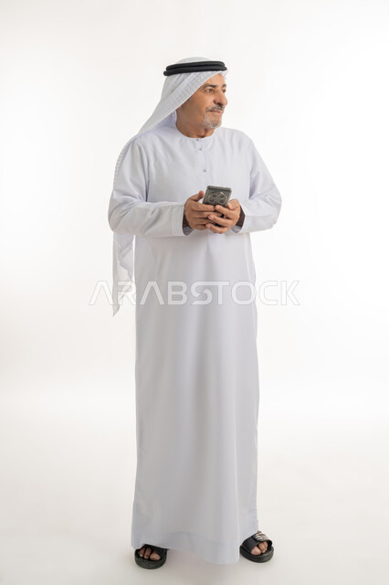 التواصل مع الافراد عبر الهاتف المحمول، دردشة ومحادثات وتعارف، بورتريه لرجل مسن عربي خليجي إماراتي مبتسم يرتدي الكندورة والغترة يستعمل الجوال، تصفح الانترنت والتسوق الالكتروني، النظر الى شيء ما بايماءات السرور، الطول الكامل للجسم، خلفية بيضاء
