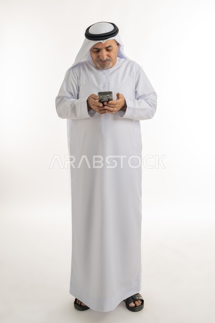 دردشة ومحادثات وتعارف، بورتريه لرجل مسن عربي خليجي إماراتي مبتسم يرتدي الكندورة والغترة يستعمل الجوال، تصفح الانترنت والتسوق الالكتروني, استخدام التقنيات والأجهزة الحديثة، التواصل مع الافراد عبر الهاتف المحمول، الطول الكامل للجسم، خلفية بيضاء