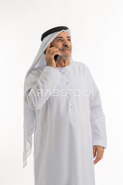 استخدام التقنيات والأجهزة الحديثة، التواصل مع الأهل والأصدقاء، إيماءات وتعابير السعادة والسرور لسماع الأخبار المفرحة، بورتريه لرجل مسن عربي خليجي إماراتي مبتسم يرتدي الكندورة والغترة يقوم بإجراء مكالمة هاتفية عبر الهاتف المحمول، خلفية بيضاء