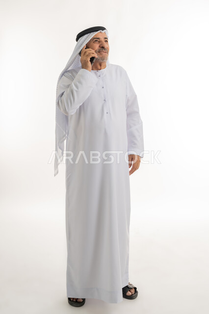 التواصل مع الأهل والأصدقاء، إيماءات وتعابير السعادة والسرور لسماع الأخبار المفرحة، بورتريه لرجل مسن عربي خليجي إماراتي مبتسم يرتدي الكندورة والغترة يقوم بإجراء مكالمة هاتفية عبر الهاتف المحمول، استخدام التقنيات والأجهزة الحديثة، الطول الكامل للجسم، خلفية بيضاء