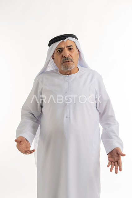 النظر للكاميرا بإيماءات الانزعاج وعدم الرضا، بورتريه لرجل عربي خليجي إماراتي كبير بالسن يرتدي الغترة والكندورة الإماراتية يرفع كفيه بتذمر وسخط، رفع اليدين في الهواء بتعابير الغضب، خلفية بيضاء