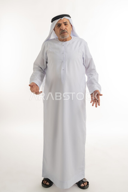 رفع اليدين في الهواء بتعابير الغضب، النظر للكاميرا بإيماءات الانزعاج وعدم الرضا، بورتريه لرجل عربي خليجي إماراتي كبير بالسن يرتدي الغترة والكندورة الإماراتية يرفع كفيه بتذمر وسخط، الجسم بالطول الكامل، خلفية بيضاء