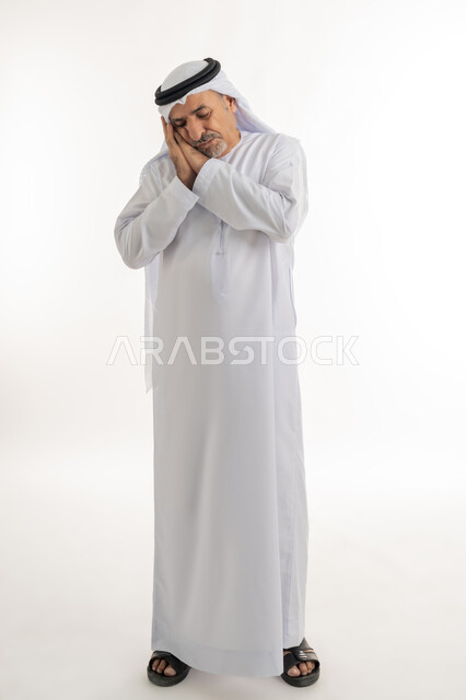 إغماض العينين والنوم بالسلام، السهر لوقت متأخر من الليل، سيد يشعر بالكسل والخمول و الارهاق، الحاجة للنوم والراحة، بورتريه لرجل مسن عربي خليجي اماراتي يرتدي الكندورة والغترة  يضع يديه تحت رأسه بإيماءات النعاس والتعب، الطول الكامل للجسم، خلفية بيضاء