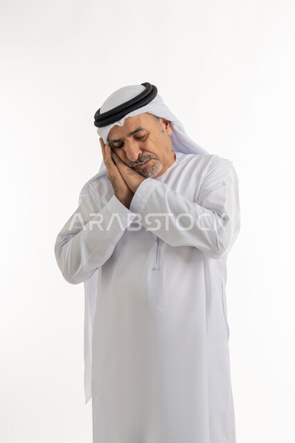الحاجة للنوم والراحة، بورتريه لرجل مسن عربي خليجي اماراتي يرتدي الكندورة والغترة  يضع يديه تحت رأسه بإيماءات النعاس والتعب، إغماض العينين والنوم بالسلام، السهر لوقت متأخر من الليل، سيد يشعر بالكسل والخمول و الارهاق، خلفية بيضاء