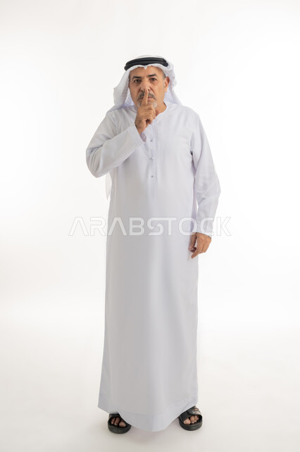 الهدوء والسكون وعدم الإزعاج، التوقف عن الكلام وخفض الصوت، بورتريه لرجل كبير السن عربي خليجي اماراتي يرتدي الكندورة و الغترة و النظارات الطبية يضع أصبع السبابة على فمه بإيماءات طلب السكوت والصمت، صورة الجسم بالطول الكامل, خلفية بيضاء