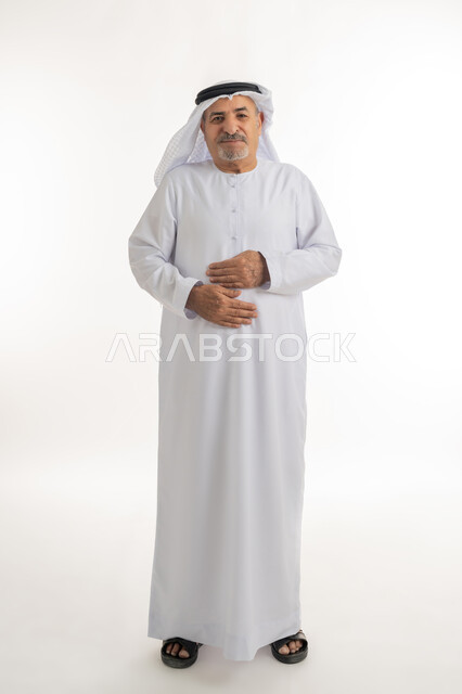 الشعور بالمرض ووجع في البطن, حركات وجه ويدين تدل على الاضطراب والانزعاج، ضرورة الراحة وأخذ العلاج المناسب، بورتريه لرجل كبير السن عربي خليجي إماراتي يرتدي الغترة والكندورة يضع يديه على بطنه بتعابير التألم, الجسم بالطول الكامل، خلفية بيضاء