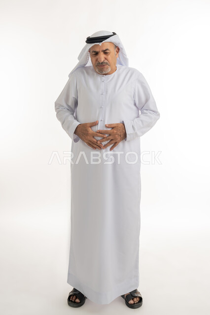 Gastrointestinal diseases and infections, need for treatment, portrait of an elderly Arab Gulf Emirati man wearing a kandura and ghutra, putting his hands on his stomach with expressions of fatigue, exhaustion and feeling sick, suffering from stomach and abdominal pain, full body image, white background