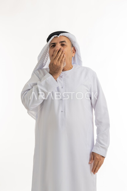 Yawning and feeling tired, lazy and lethargic, feeling tired and needing to sleep and rest, portrait of an elderly Arab Gulf Emirati man wearing a kandura and ghutra, putting his hand on his mouth with gestures indicating drowsiness, white background