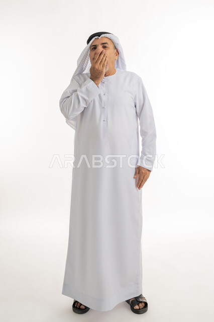 الإحساس بالإرهاق و الحاجه الى النوم والراحه ,بورتريه لرجل كبير السن عربي خليجي اماراتي يرتدي الكندورة والغترة يضع يده على فمه بإيماءات تدل على النعاس, التثاؤب والشعور بالتعب و الكسل والخمول، الطول الكامل للجسم, خلفية بيضاء
