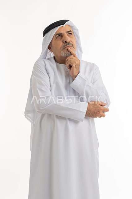 النظر إلى مكان بعيد بشرود, بورتريه لرجل كبير بالسن عربي خليجي اماراتي يرتدي الكندورة والغترة يقف ويضع يده على ذقنه بإيماءات القلق والتفكير بشيء ما, التخطيط للمستقبل ووضع الأهداف المستقبلية, خلفية بيضاء