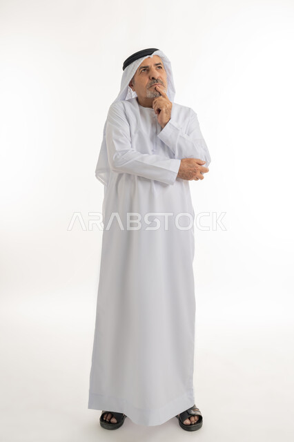 Planning for the future and setting future goals, looking away absentmindedly, portrait of an elderly Arab Gulf Emirati man wearing a kandura and ghutra standing and putting his hand on his chin with gestures of worry and thinking about something, full body image, white background