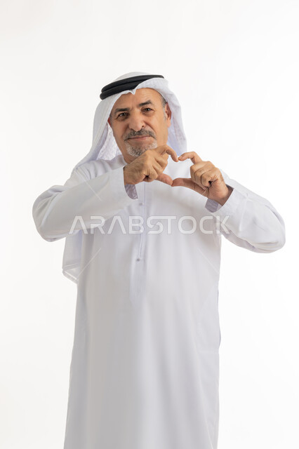 مفهوم المحبة والمودة، ايماءات يدين على شكل قلب،, بورتريه لرجل عربي خليجي سعودي مبتسم يرتدي الكندورة والغترة ينظر إلى الكاميرا  بتعابير السعادة والفرح,  اظهار مشاعر الحب والشغف, خلفية بيضاء