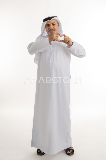 اظهار مشاعر الحب والشغف، مفهوم المحبة والمودة، ايماءات يدين على شكل قلب،, بورتريه لرجل عربي خليجي سعودي مبتسم يرتدي الكندورة والغترة ينظر إلى الكاميرا  بتعابير السعادة والفرح, صورة الجسم بالطول الكامل, خلفية بيضاء
