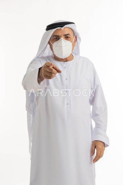 حركات يدين تدل على هذا المكان (هنا)، تجنب الملامسه السطحيه والحفاظ على المسافات، إتباع الإجراءات الوقائيه ضد فايروس كورونا، بورتريه لرجل مسن عربي خليجي اماراتي يرتدي الكندورة والغتره يضع القناع الطبي يشير باصبعه الى الامام، تحديد موقع شيء معين، خلفيه بيضاء