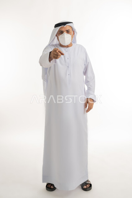 Locating a specific object, avoiding surface contact and maintaining distances, following preventive measures against the Corona virus, portrait of an elderly Arab Gulf Emirati man wearing a kandura and a ghutra, wearing a medical mask, pointing his thumb forward, full body length, white background