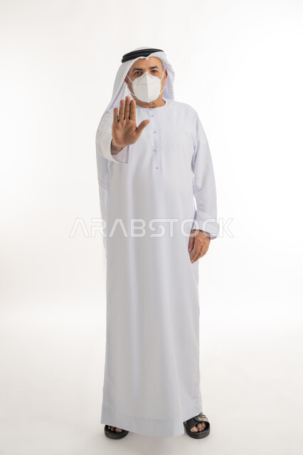 إيماءات التوقف وعدم الاقتراب، الالتزام بإجراءات الوقاية، الحفاظ على المسافات وعدم الملامسة السطحية, تجنب انتشار العدوى بكوفيد 19، بورتريه لرجل عربي خليجي اماراتي مسن يرتدي الكندورة والغترة والكمامة الواقية ينظر الى الكاميرا يرفع يده للاعلى، بخلفية بيضاء