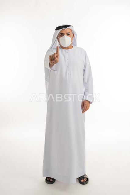 اتباع الإجراءات الاحترازية لمنع انتشار العدوى، بورتريه لرجل كبير بالسن عربي خليجي اماراتي يرتدي الكندورة والغترة يضع الكمامة الطبية على وجهه ويرفع اصبع السبابة نحو الأعلى، اتخاذ التدابير اللازمة للوقاية من فيروس كورونا، الجسم بالطول الكامل، خلفية بيضاء