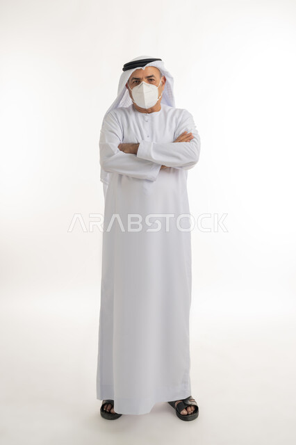 تجنب انتشار العدوى بكوفيد 19، بورتريه لرجل مسن عربي خليجي اماراتي يرتدي الكندورة والغترة والكمامة الواقية ينظر الى الكامير ويقف بايماءات ايدي متقاطعة، الالتزام بإجراءات الوقاية، صورة الجسم بالطول الكامل، خلفية بيضاء