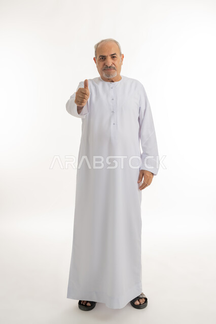 تعابير القبول والفرح والسرور، الاهتمام بالأناقة والمظهر الخارجي، بورتريه لرجل كبير بالسن عربي خليجي إماراتي يرتدي الكندورة ينظر الى الكاميرا يرفع إصبع السبابة للاعلى بإيماءات الإعجاب والرضا، صورة الجسم بالطول الكامل، خلفية بيضاء