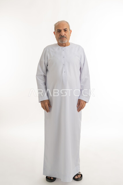 الرجولة والأناقه والاهتمام بالمظهر الخارجي، النظر إلى الكاميرا يإيماءات الجدية والحزم، بورتريه لرجل كبير بالسن عربي خليجي اماراتي يرتدي الكندورة يقف باستقامة بتعابير الثقة بالنفس, صورة الجسم بالطول الكامل، خلفية بيضاء