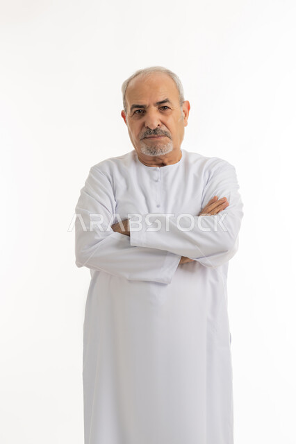 مفهوم الاهتمام بالمظهر الخارجي و الرجولة والأناقه، بورتريه لرجل كبير بالسن عربي خليجي اماراتي يرتدي الكندورة يقف بإيدي متقاطعه بتعابير الثقة بالنفس, النظر إلى الكاميرا يإيماءات الجدية والحزم، خلفية بيضاء