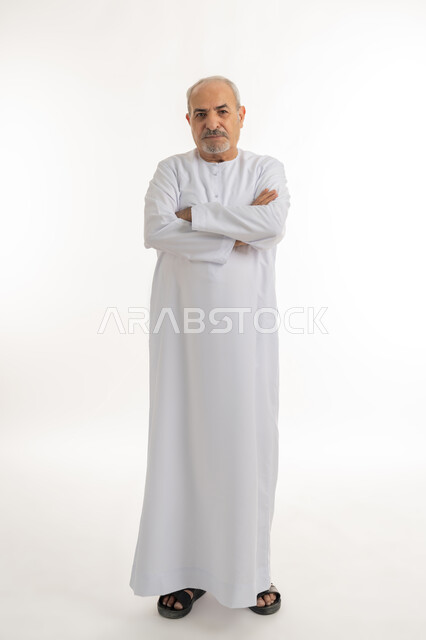 الوقوف باستقامة بايدي متقاطعه بايماءات الثقة بالنفس، مفهوم الاهتمام بالمظهر الخارجي و الرجولة والأناقه، بورتريه لرجل مسن عربي خليجي اماراتي يرتدي الكندورة, النظر إلى الكاميرا بتعابيرالجدية والحزم، صورة الجسم بالطول الكامل، خلفية بيضاء