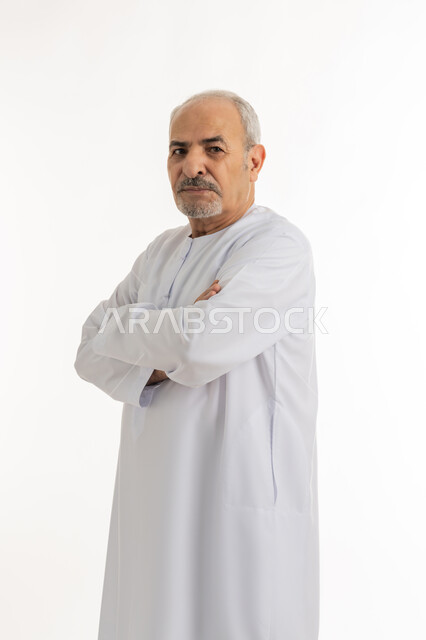 النظر إلى الكاميرا يإيماءات الجدية والحزم، مفهوم الاهتمام بالمظهر الخارجي و الرجولة والأناقه، بورتريه لرجل كبير بالسن عربي خليجي اماراتي يرتدي الكندورة يقف بإيدي متقاطعه بتعابير الثقة بالنفس, خلفية بيضاء