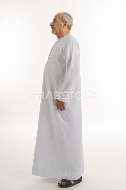 إيماءات الجدية والحزم، التقدم بالعمر والهيبة والوقار، بورتريه من الجانب لرجل كبير بالسن عربي خليجي إماراتي يرتدي الكندورة ينظر الى شيء ما, الاهتمام بالأناقة والمظهر الخارجي، الوقوف باستقامة وثبات، مفهوم الرجولة العربية, الطول الكامل للجسم، خلفية بيضاء