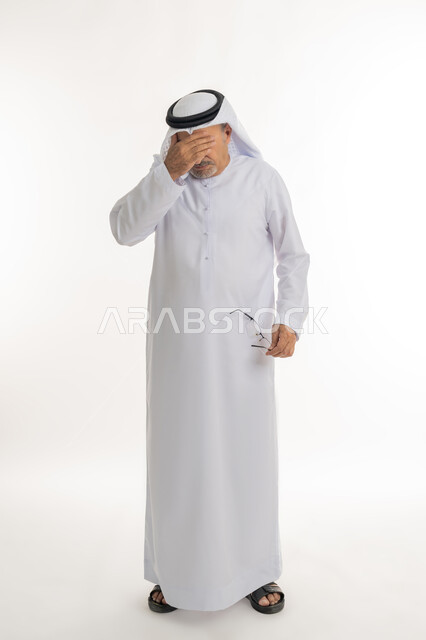 Putting hand on face and pressing eyes due to severe pain, suddenly remembering something specific, portrait of an elderly Arab Gulf Emirati man wearing traditional kandura and ghutra holding his glasses in his hand, suffering from vision problems due to old age, full body image, white background