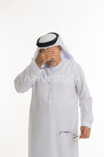 المعاناة من مشاكل في النظر بسبب تقدم العمر, وضع اليد على الوجه والضغط على العيون من شدة الوجع, تذكر شيء محدد بشكل مفاجئ, بورتريه لرجل كبير بالسن عربي خليجي اماراتي يرتدي الكندورة التقليدية والغترة يمسك نظارته الطبية بيده، خلفية بيضاء