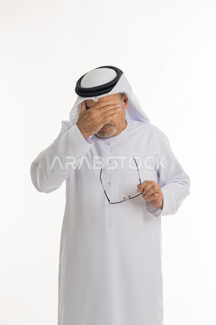 تذكر شيء محدد بشكل مفاجئ, بورتريه لرجل كبير بالسن عربي خليجي اماراتي يرتدي الكندورة التقليدية والغترة يمسك نظارته الطبية بيده، المعاناة من مشاكل في النظر بسبب تقدم العمر, وضع اليد على الوجه والضغط على العيون من شدة الوجع, خلفية بيضاء