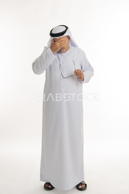 وضع اليد على الوجه والضغط على العيون من شدة الوجع, تذكر شيء محدد بشكل مفاجئ, بورتريه لرجل كبير بالسن عربي خليجي اماراتي يرتدي الكندورة التقليدية والغترة يمسك نظارته الطبية بيده، المعاناة من مشاكل في النظر بسبب تقدم العمر, صورة الجسم بالكامل، خلفية بيضاء