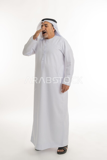 التثاؤب والشعور بالتعب و الكسل والخمول، الإحساس بالإرهاق و الحاجه الى النوم والراحه ,بورتريه لرجل كبير السن عربي خليجي اماراتي يرتدي الكندورة والغترة يضع يده على فمه بإيماءات تدل على النعاس, الطول الكامل للجسم, خلفية بيضاء