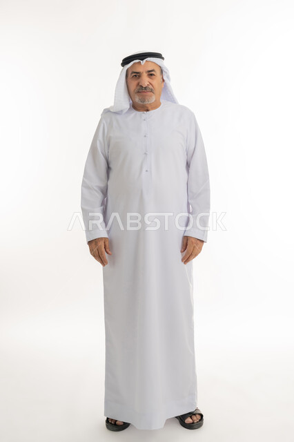 مفهوم الاهتمام بالمظهر الخارجي و الرجولة والأناقه، النظر إلى الكاميرا يإيماءات الجدية والحزم، بورتريه لرجل كبير بالسن عربي خليجي اماراتي يرتدي الكندورة و الغترة يقف باستقامة بتعابير الثقة بالنفس, صورة الجسم بالطول الكامل، خلفية بيضاء