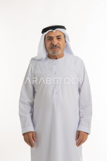 النظر إلى الكاميرا يإيماءات الجدية والحزم، بورتريه لرجل كبير بالسن عربي خليجي اماراتي يرتدي الكندورة و الغترة يقف باستقامة بتعابير الثقة بالنفس, مفهوم الاهتمام بالمظهر الخارجي و الرجولة والأناقه، خلفية بيضاء