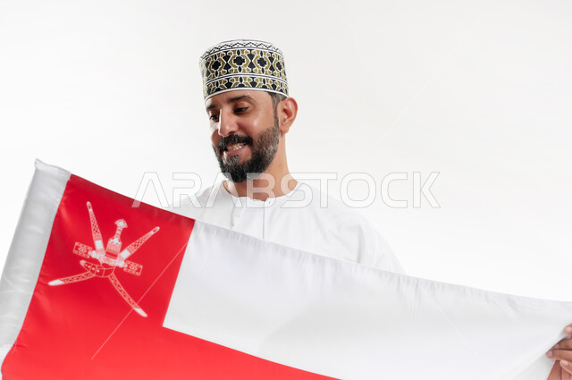 الفخر وإعتزاز بالهوية الوطنية، حب الوطن والانتماء إليه، إحياء ذكرى تأسيس الدولة 1775 م، العلم الوطني للبلاد، بورتريه  لرجل عربي خليجي عماني يرتدي الدشداشة و الكمة العمانية التقليدية يحتفل بذكرى اليوم الوطني العماني 18 نوفمبر، خلفية بيضاء